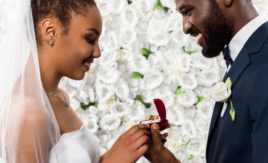 cheerful african american bride looking at box with ring near happy bridegroom and flowers